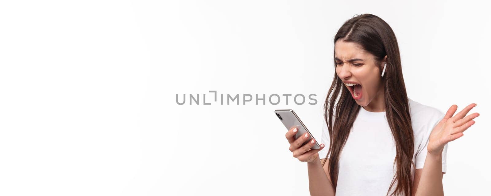 Close-up portrait of pissed-off aggressive and angry young brunette woman yelling at person while having phone call in wireless earphones, screaming at smartphone outraged.