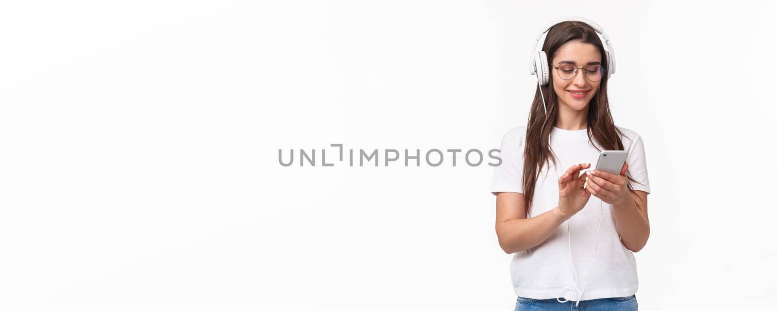 Technology, lifestyle and music concept. Portrait of lovely young feminine girl in headphones, texting friend mobile phone, cute smiling at screen smartphone, choosing podcast.