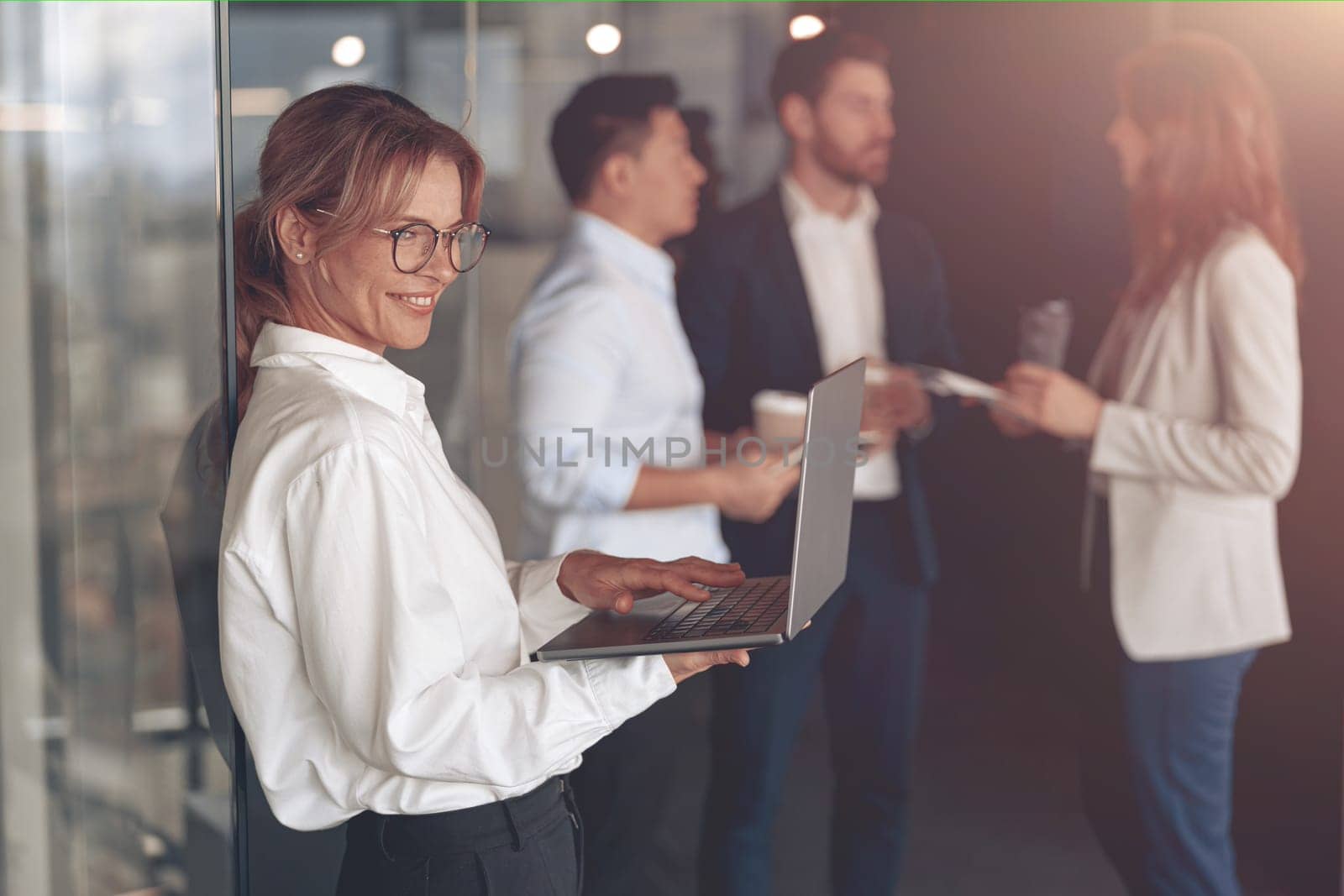 Mature smiling businesswoman at office with group of colleagues on background, working on laptop