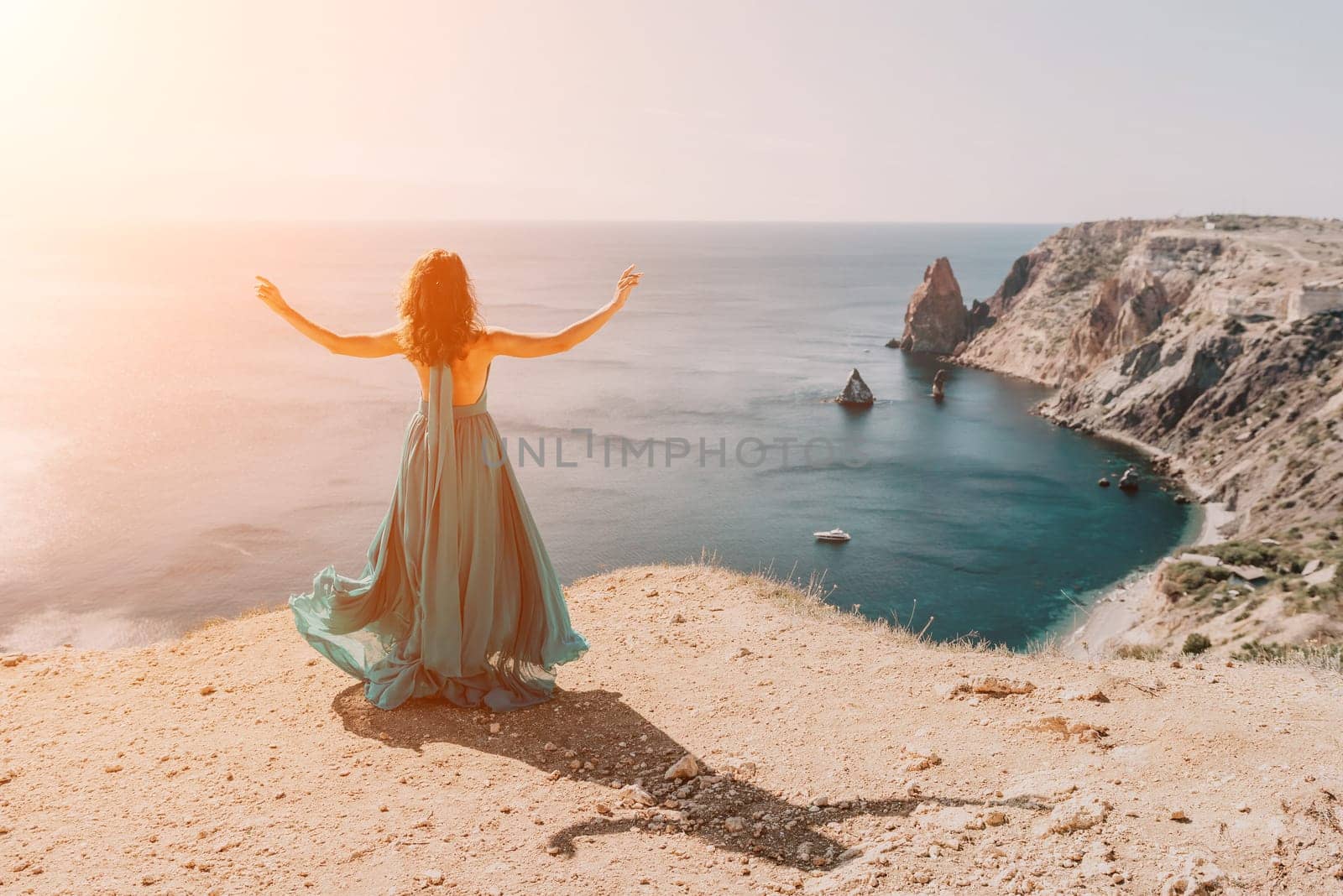 Side view a Young beautiful sensual woman in a red long dress posing on a rock high above the sea during sunrise. Girl on the nature on blue sky background. Fashion photo.