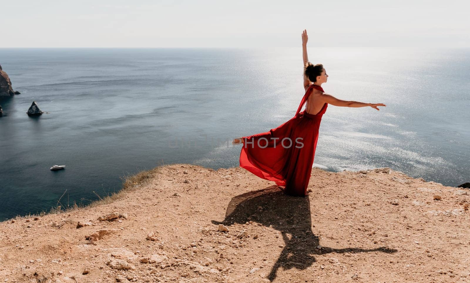 Woman in red dress on sea. Side view a Young beautiful sensual woman in a red long dress posing on a rock high above the sea on sunset. Girl on the nature on blue sky background. Fashion photo. by panophotograph