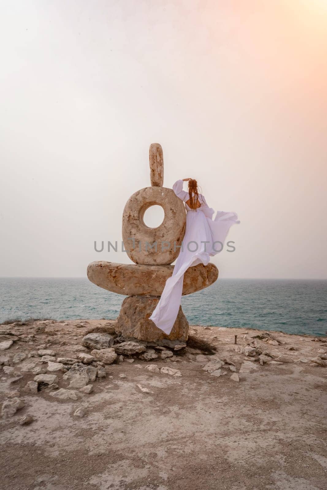 Woman sea stone. A woman stands on a stone sculpture made of large stones. She is dressed in a white long dress, against the backdrop of the sea and sky. The dress develops in the wind. by Matiunina