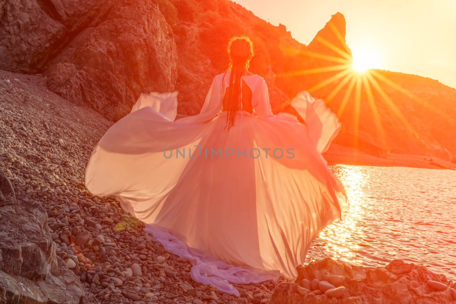 Mysterious woman silhouette long hair walks on the beach ocean water, sea nymph wind listens to the wave. Throws up a long white dress, a divine sunset. Artistic photo from the back without a face by Matiunina