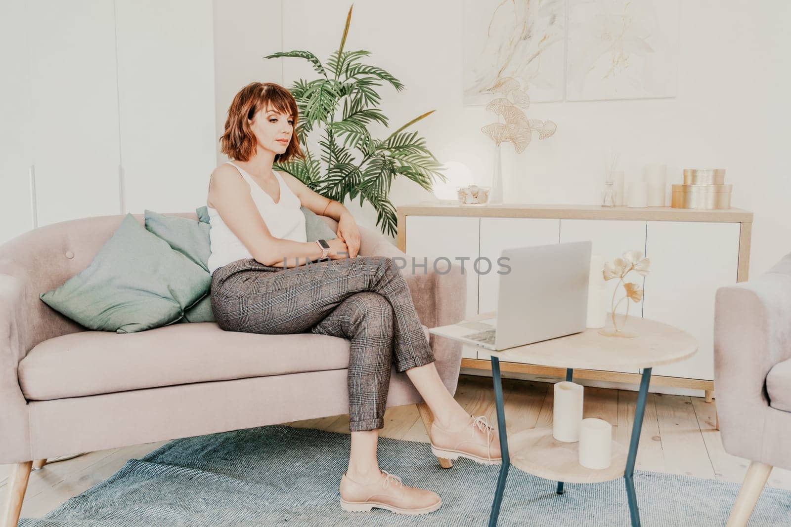 Brunette macbook sofa. Attractive young woman working on a laptop while sitting on the couch at home. She is wearing a light blouse and trousers. by Matiunina