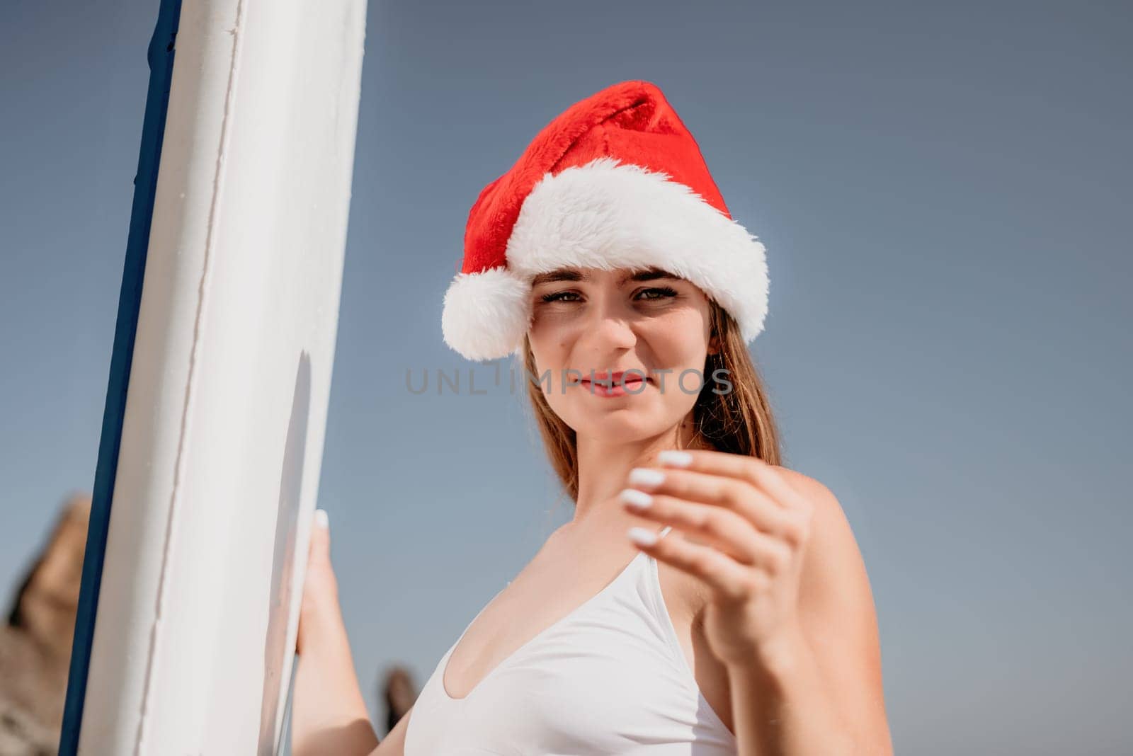 Woman sea sup. Close up portrait of happy young caucasian woman with long hair in Santa hat looking at camera and smiling. Cute woman portrait in a white bikini posing on sup board in the sea by panophotograph