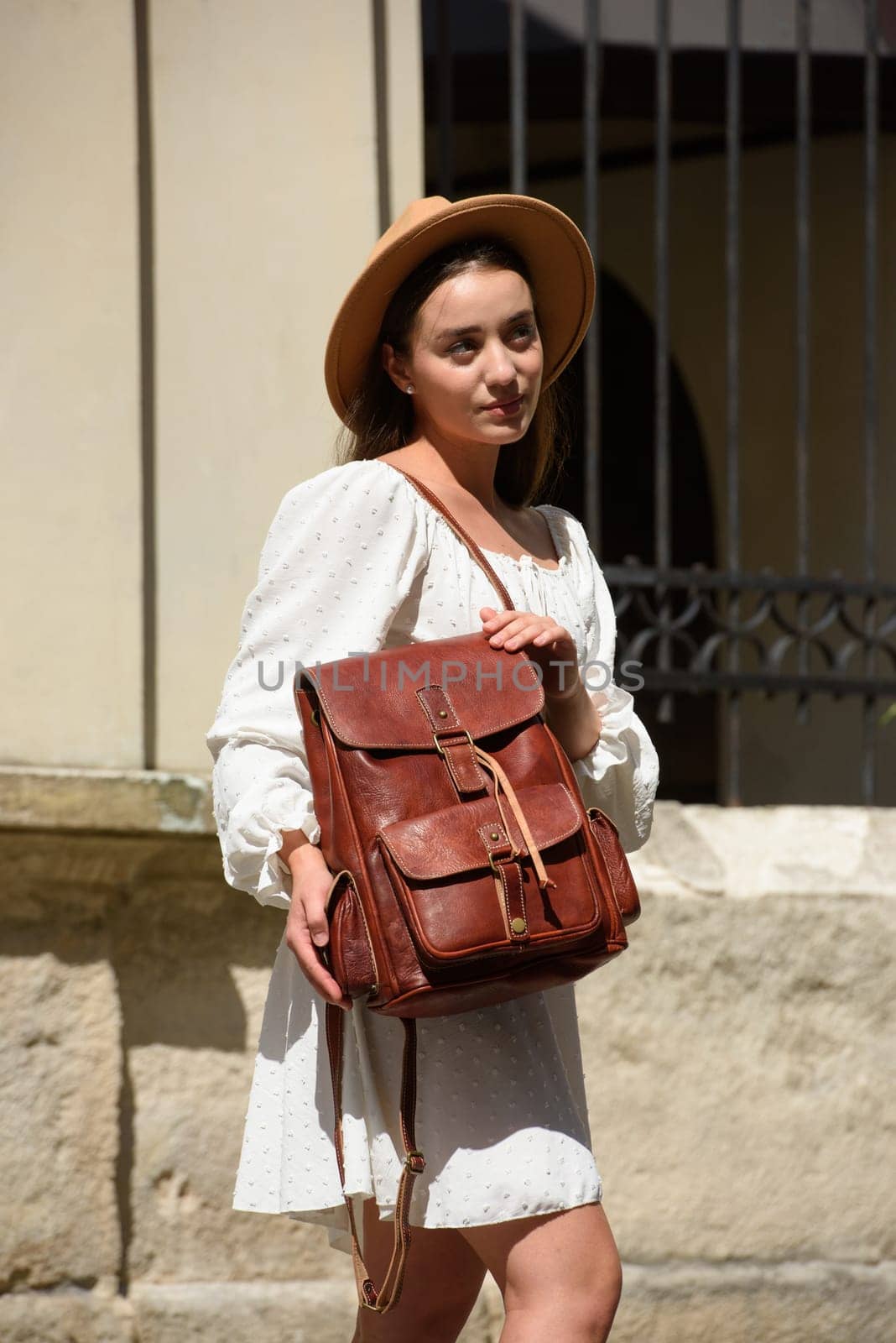 brunette with long hair wearing white dress and beige hat posing with luxury leather backpack. old city street. Vacation, tourism and travel