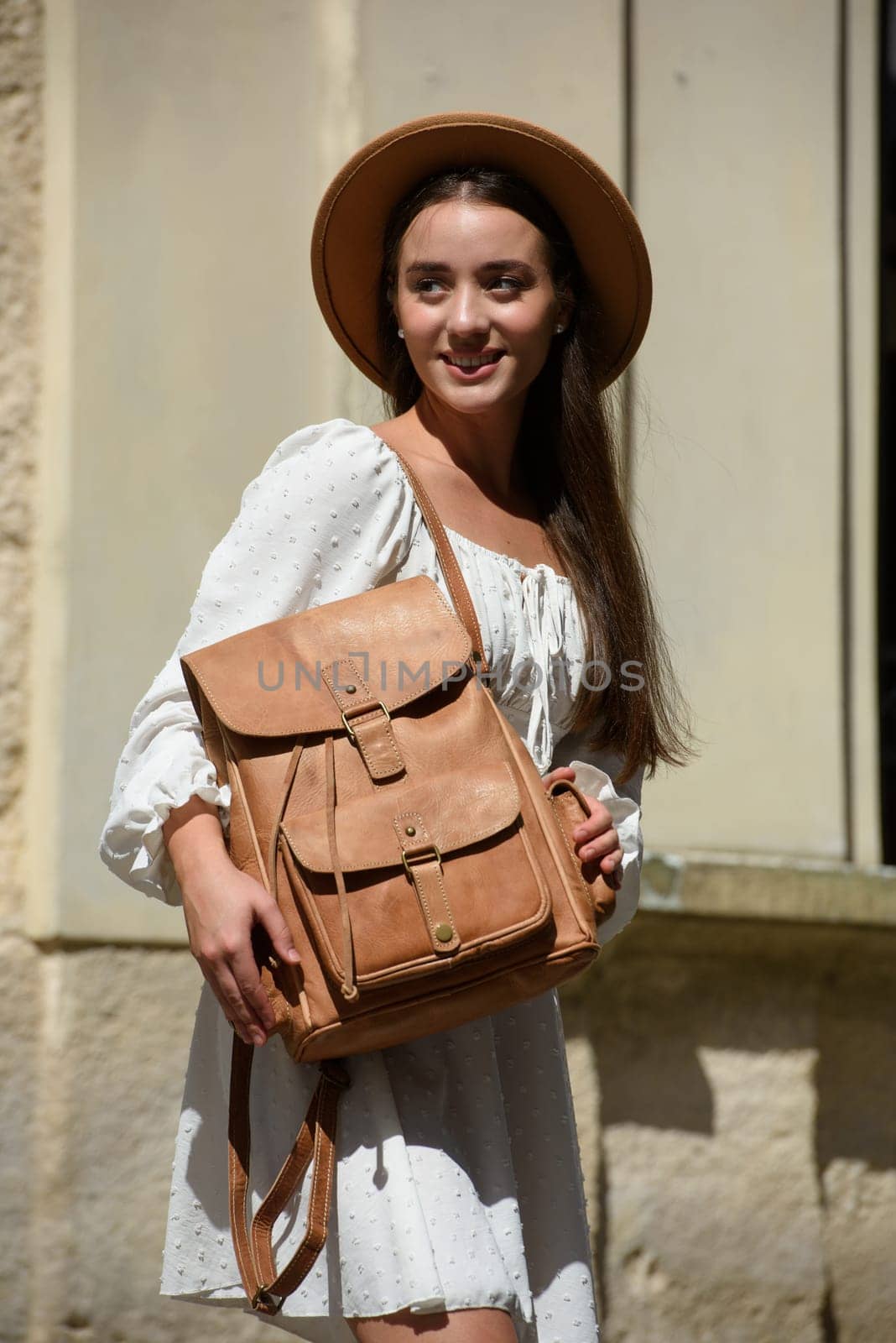 brunette with long hair wearing white dress and beige hat posing with luxury leather backpack. old city street. Vacation, tourism and travel