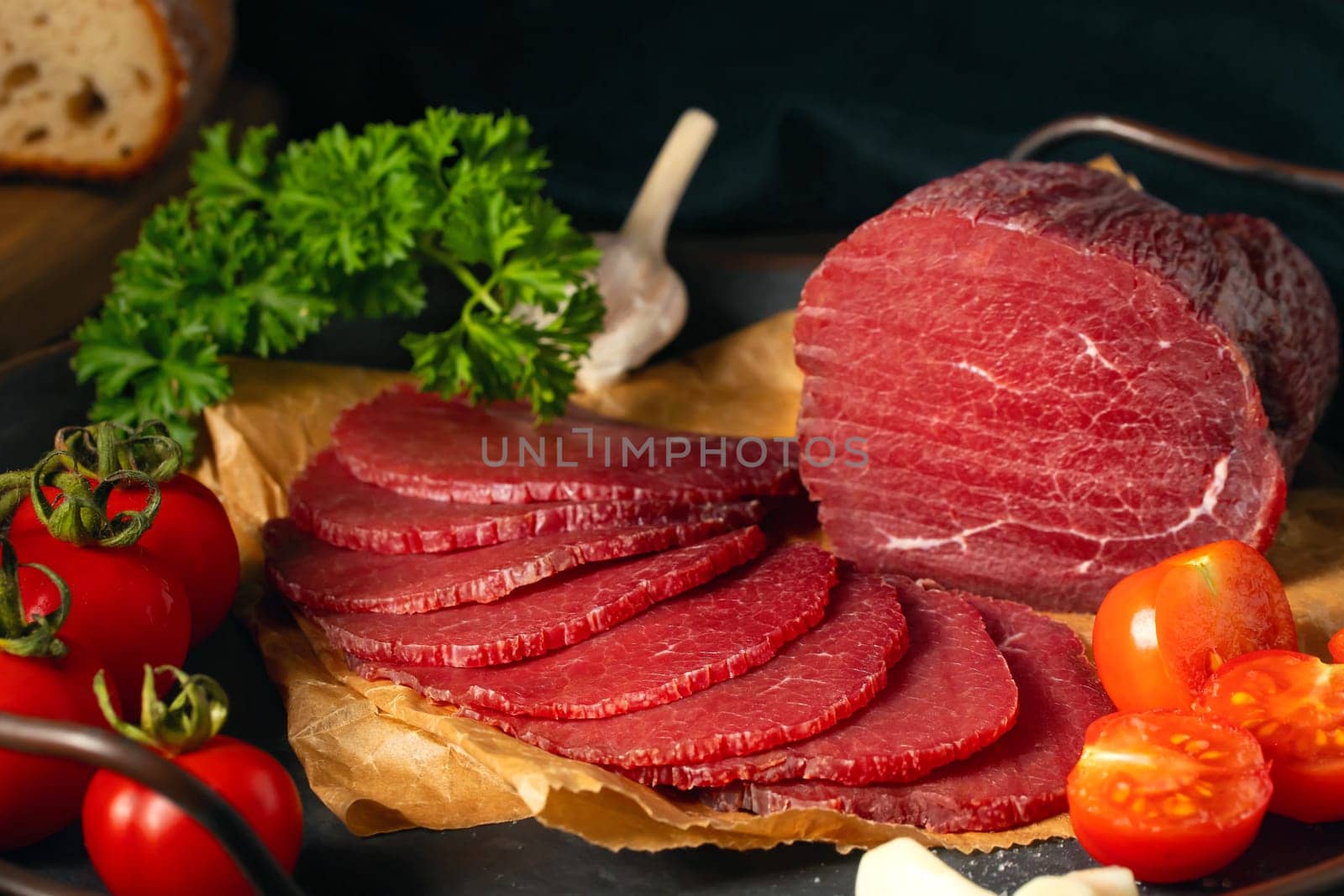 Whole and sliced bresaola on a metal round tray with tomatoes, garlic and parsley by galsand
