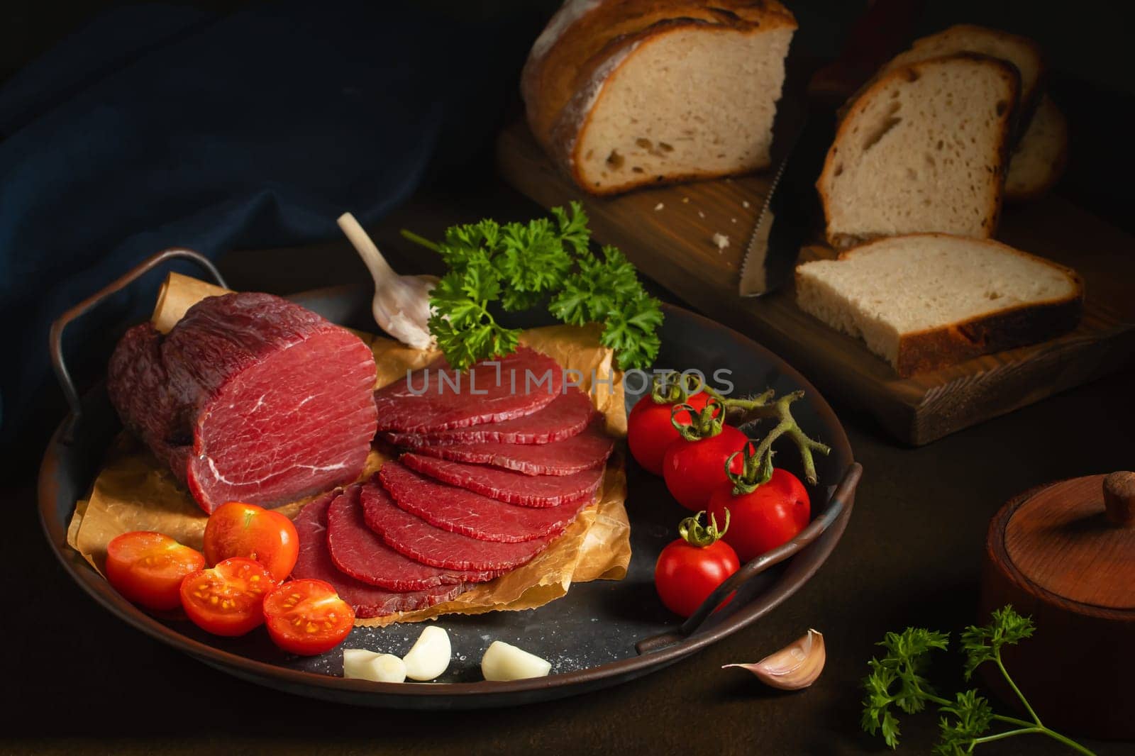 Whole and sliced bresaola on a metal round tray with tomatoes, garlic and bread by galsand