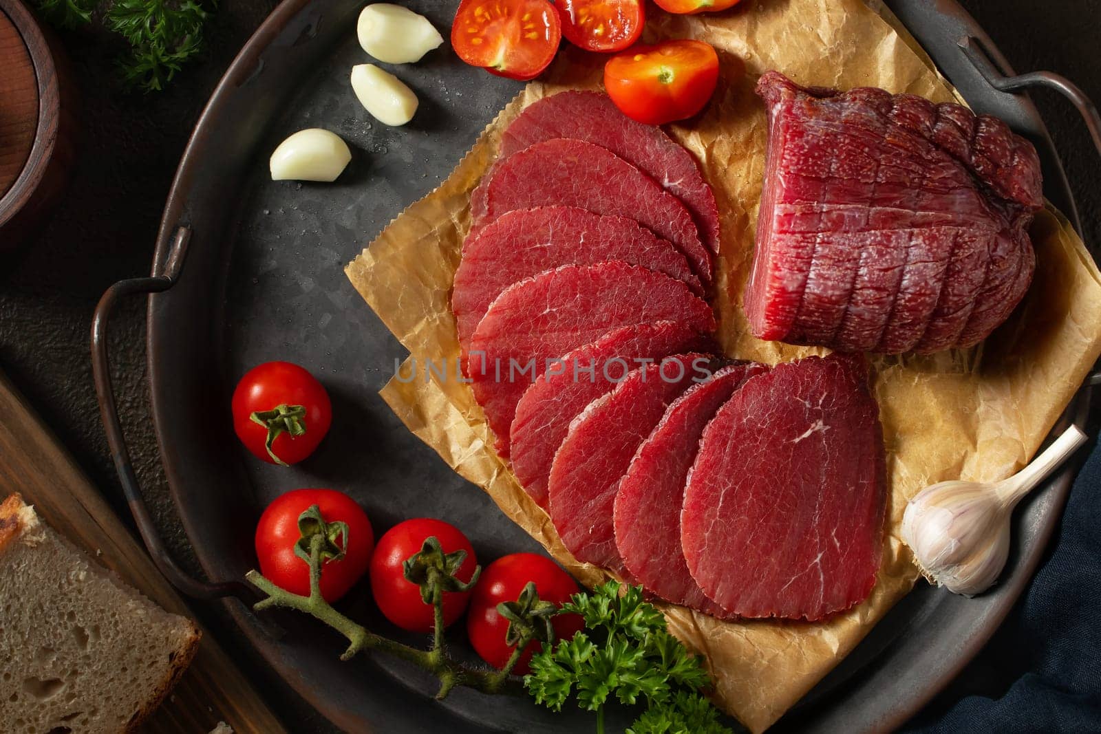 Whole and sliced bresaola on a metal round tray with tomatoes, garlic and bread by galsand