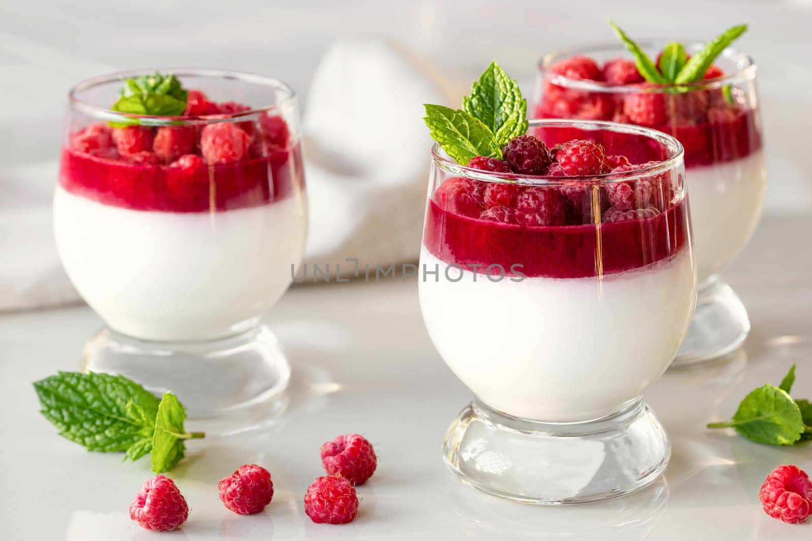 Panna cotta with raspberry jelly and mint leaves in glass glasses on a white table by galsand
