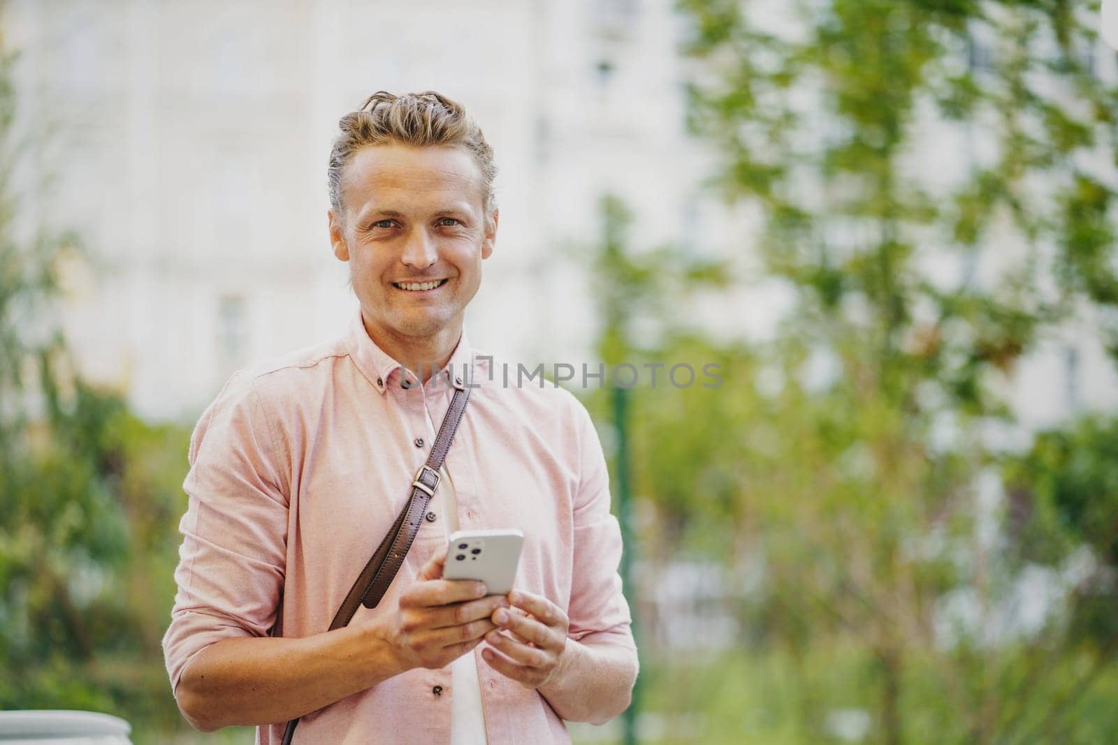 Happy man, satisfied mobile phone customer, smiling on city street. Engaged in mobile communication, he texts message or uses mobile app, reflecting seamless integration of technology into daily life by LipikStockMedia