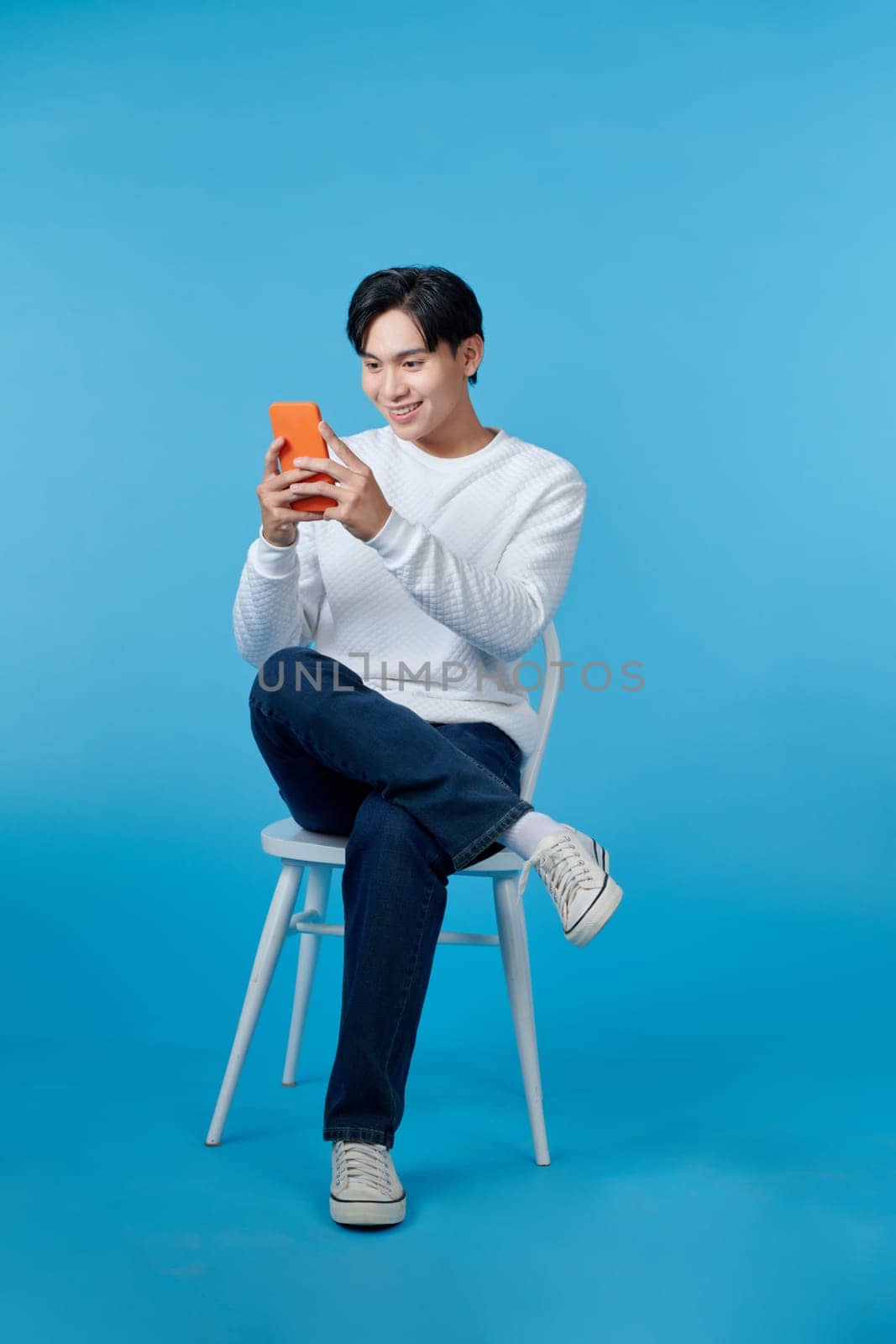 Young Asian man sitting and using smartphone on blue background by makidotvn