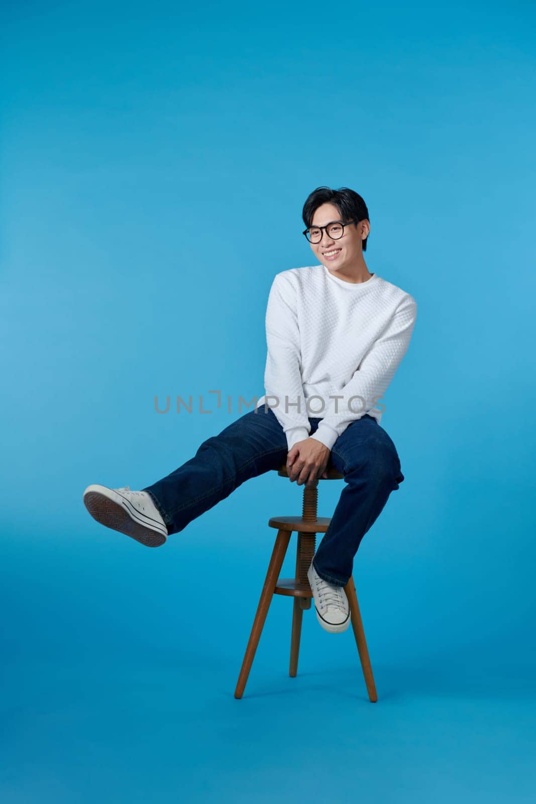 Vertical shot of a young cheerful man sitting on a stool isolated on blue background
