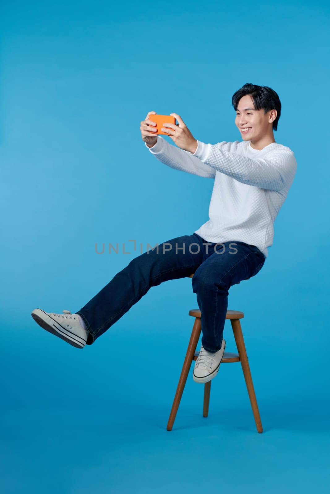 Full body of young man sit a stool playing game cellphone isolated over blue background by makidotvn