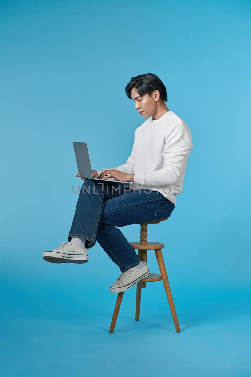 Handsome emotional man sitting on stool and using laptop isolated on blue background