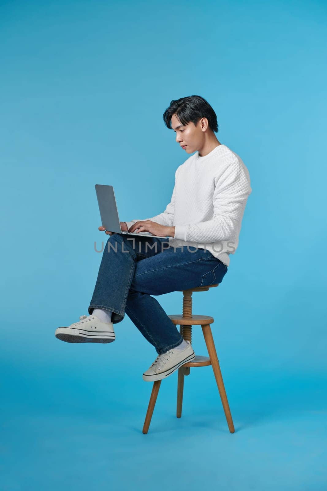 Young Asian man sitting and using laptop on bluebackground by makidotvn