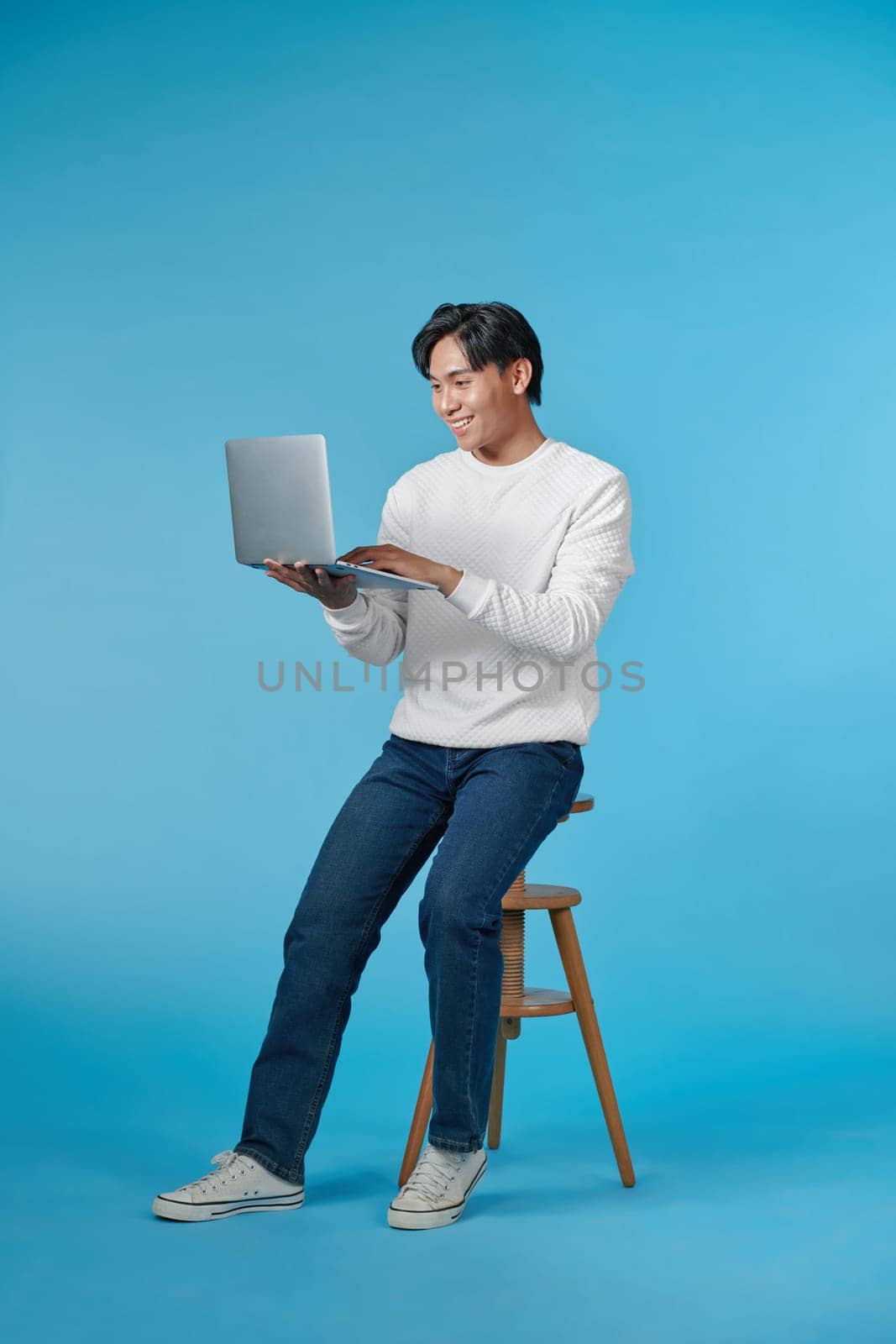 Young Asian man sitting and using laptop on bluebackground by makidotvn
