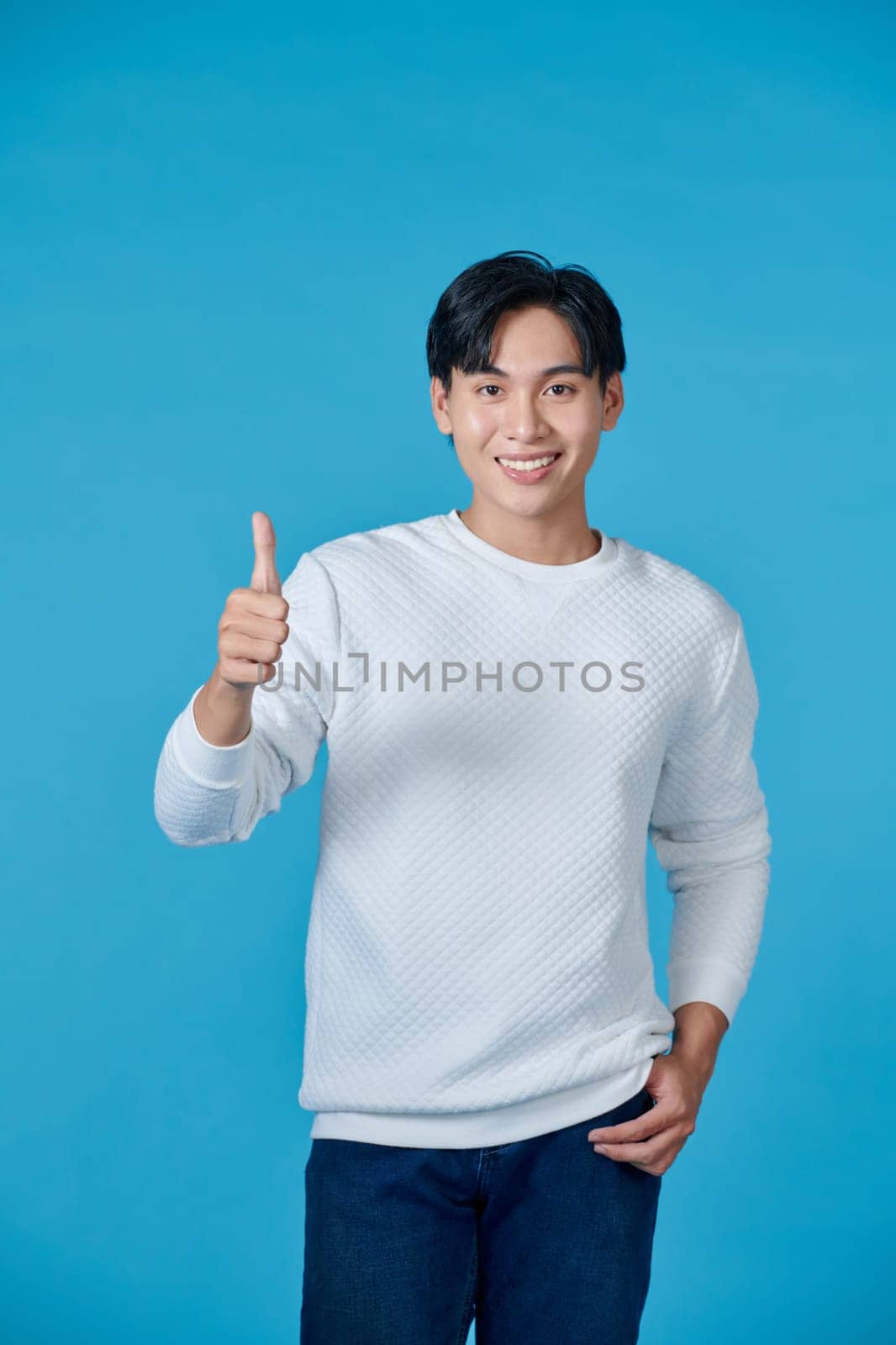 Smiling young asian man wearing white jumper and jeans on blue background giving thumbs up by makidotvn