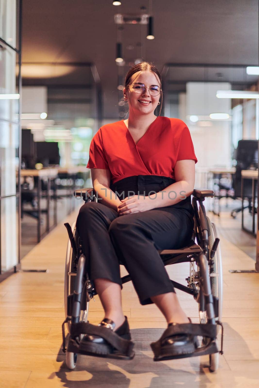 A modern young businesswoman in a wheelchair is surrounded by an inclusive workspace with glass-walled offices, embodying determination and innovation in the business world.