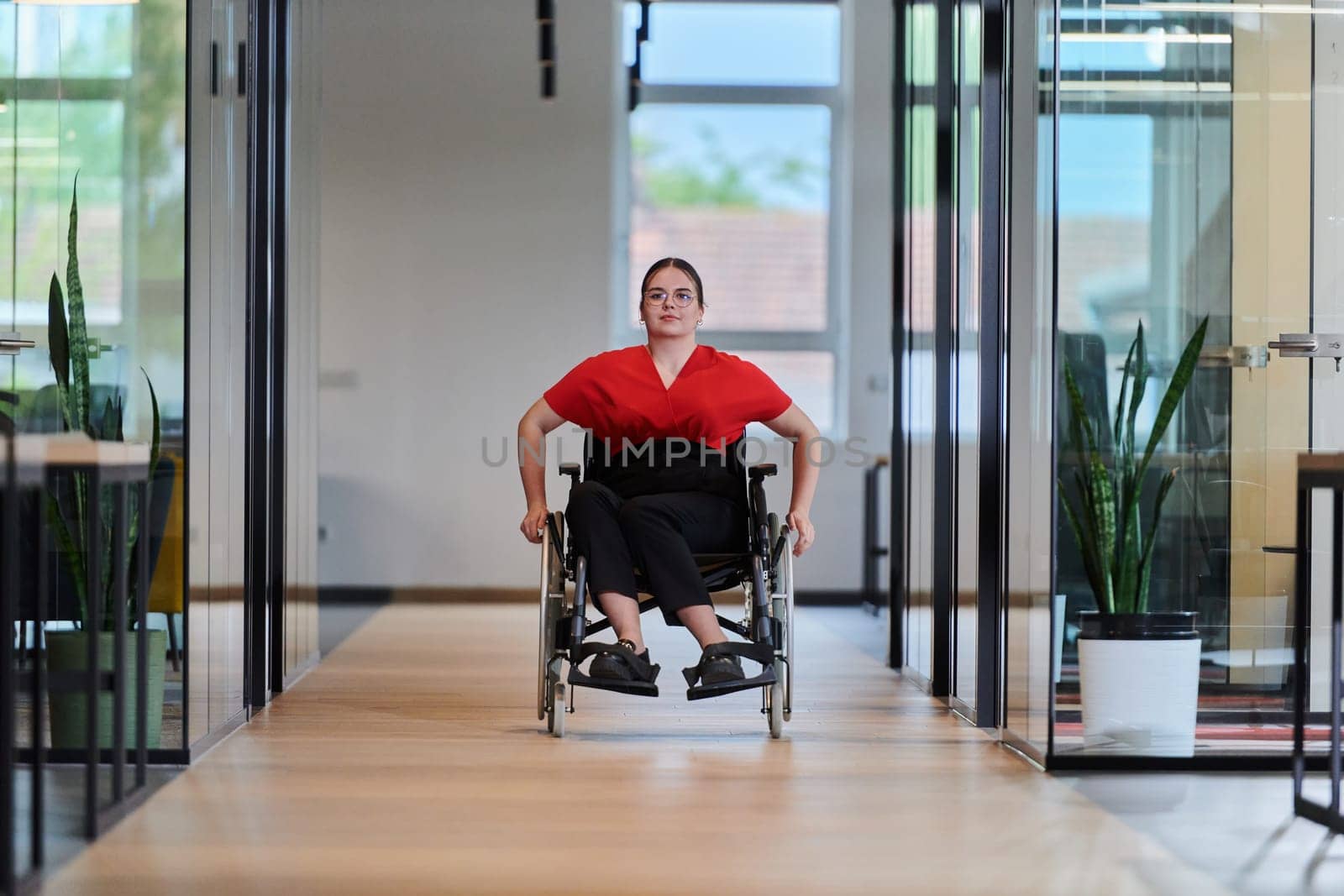 A modern young businesswoman in a wheelchair is surrounded by an inclusive workspace with glass-walled offices, embodying determination and innovation in the business world.