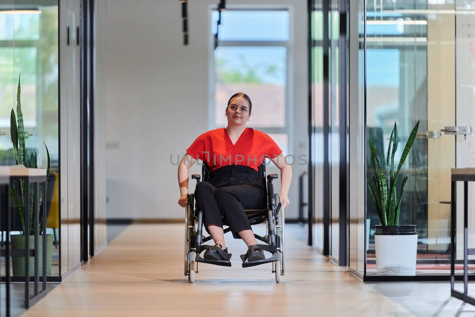 A modern young businesswoman in a wheelchair is surrounded by an inclusive workspace with glass-walled offices, embodying determination and innovation in the business world.