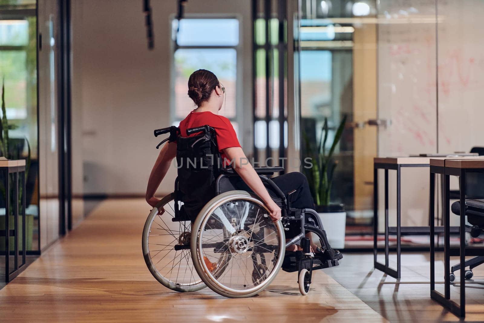 A modern young businesswoman in a wheelchair is surrounded by an inclusive workspace with glass-walled offices, embodying determination and innovation in the business world by dotshock