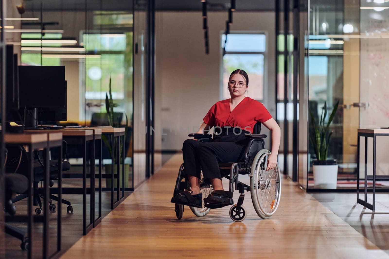 A modern young businesswoman in a wheelchair is surrounded by an inclusive workspace with glass-walled offices, embodying determination and innovation in the business world.