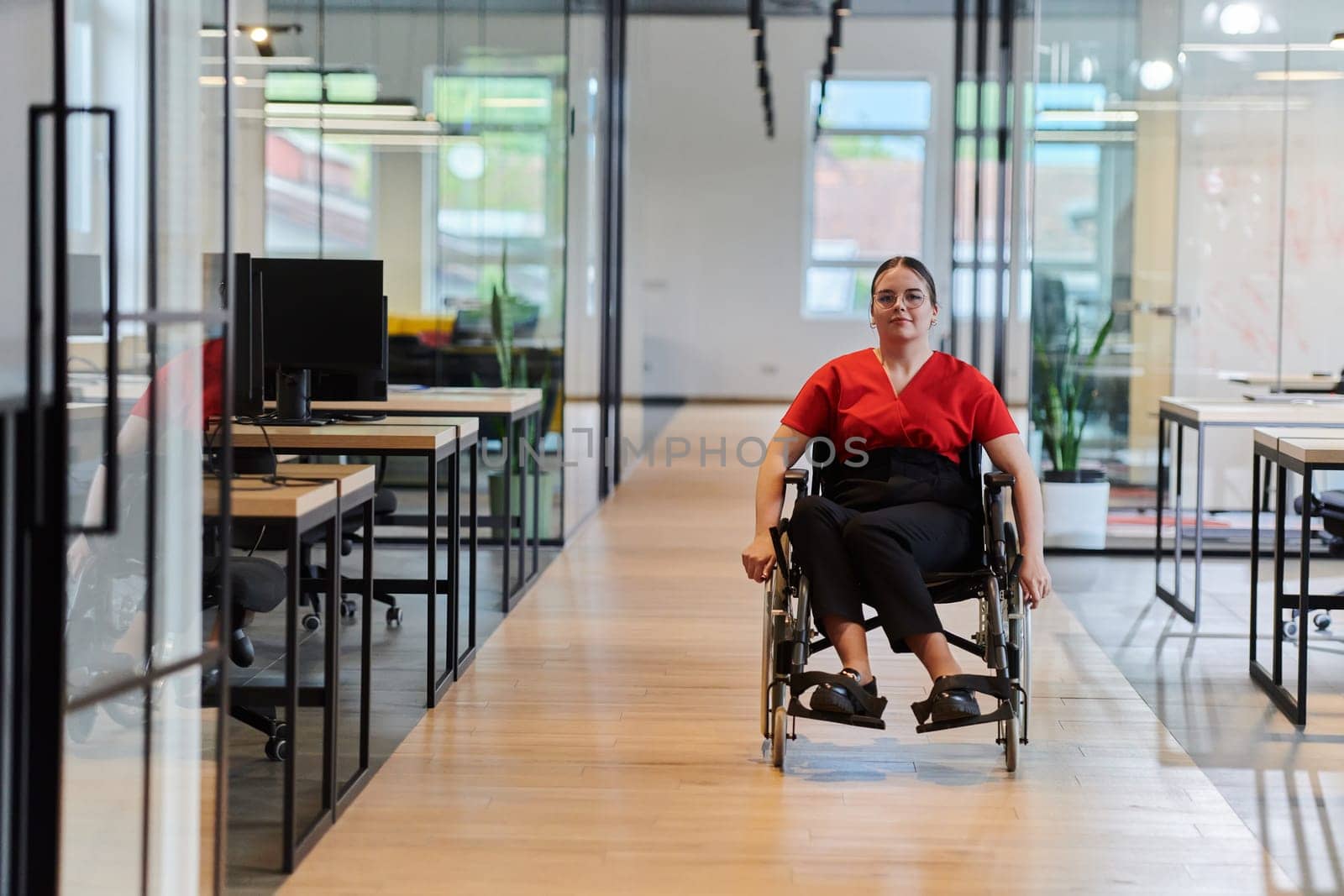 A modern young businesswoman in a wheelchair is surrounded by an inclusive workspace with glass-walled offices, embodying determination and innovation in the business world.