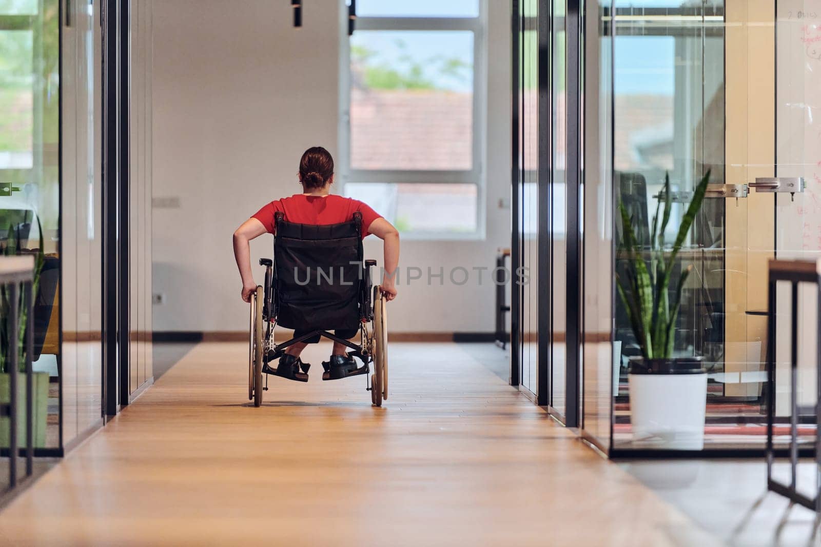 A modern young businesswoman in a wheelchair is surrounded by an inclusive workspace with glass-walled offices, embodying determination and innovation in the business world.