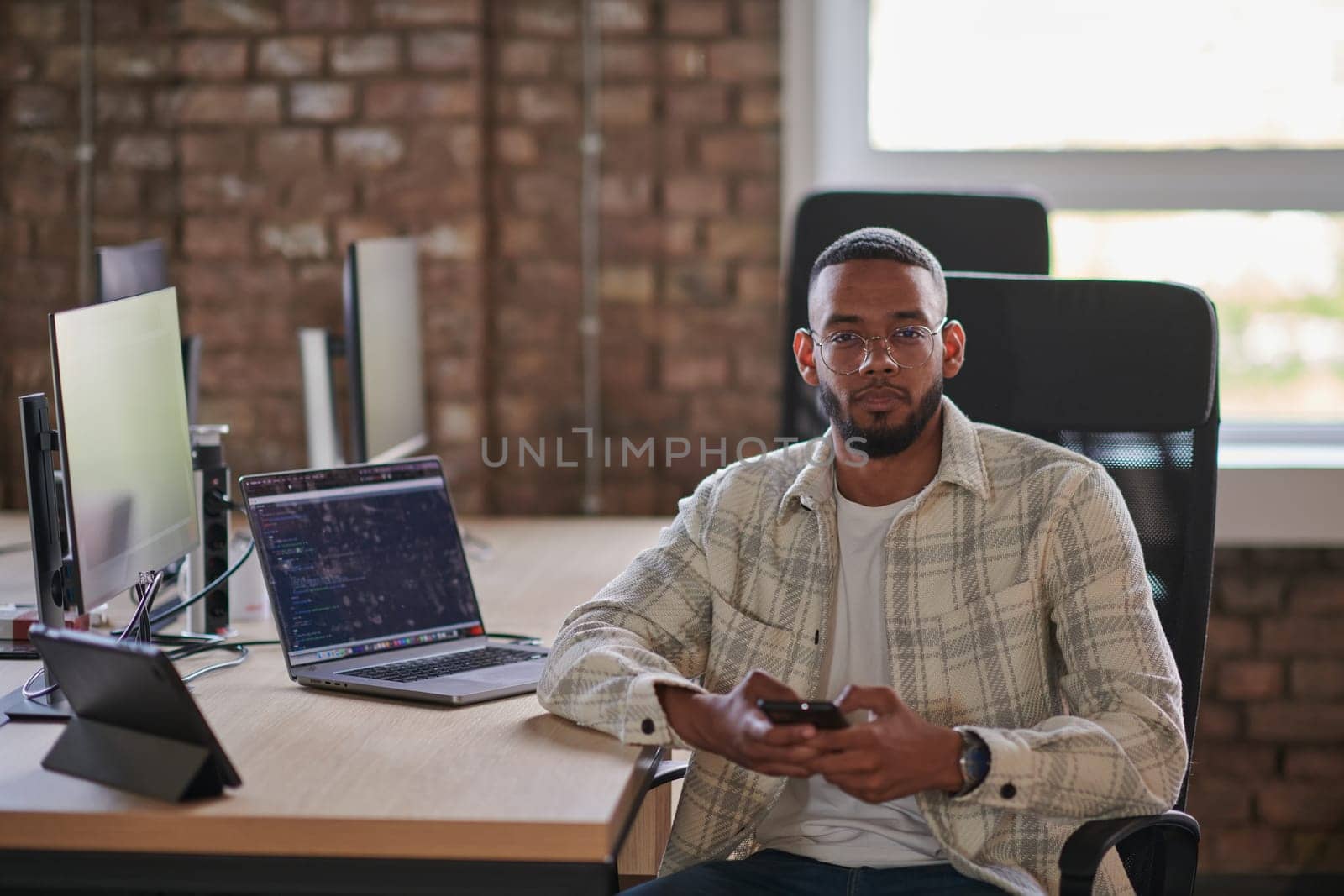 A young African American entrepreneur takes a break in a modern office, using a smartphone to browse social media, capturing a moment of digital connectivity and relaxation amidst his business endeavors. by dotshock