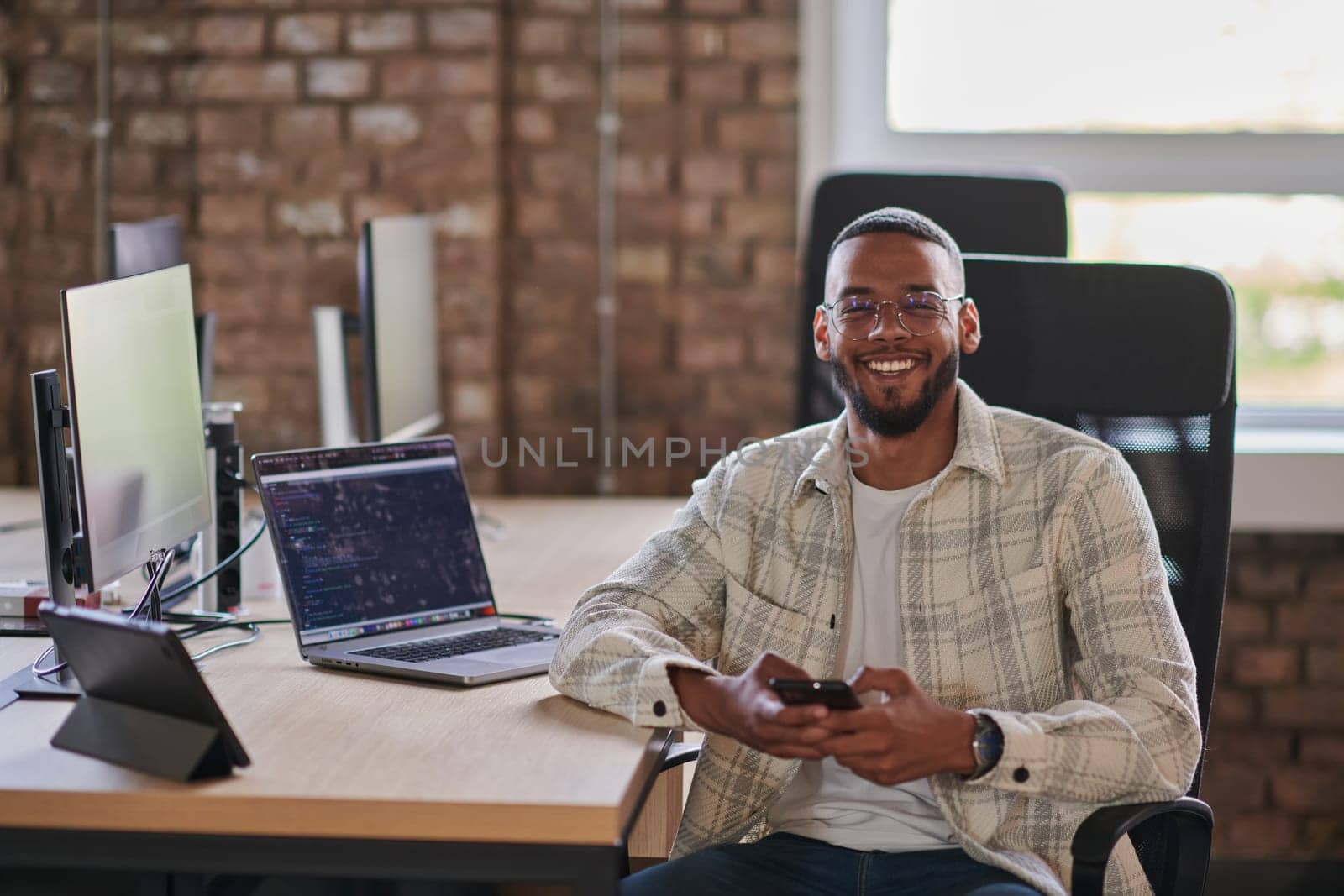 A young African American entrepreneur takes a break in a modern office, using a smartphone to browse social media, capturing a moment of digital connectivity and relaxation amidst his business endeavors. by dotshock