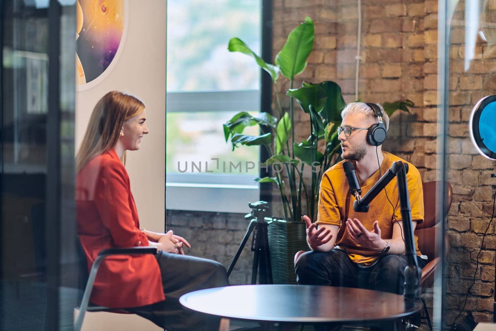 A gathering of young business professionals, some seated in a glass-walled office, engage in a lively conversation and record an online podcast, embodying modern collaboration and dynamic interaction.