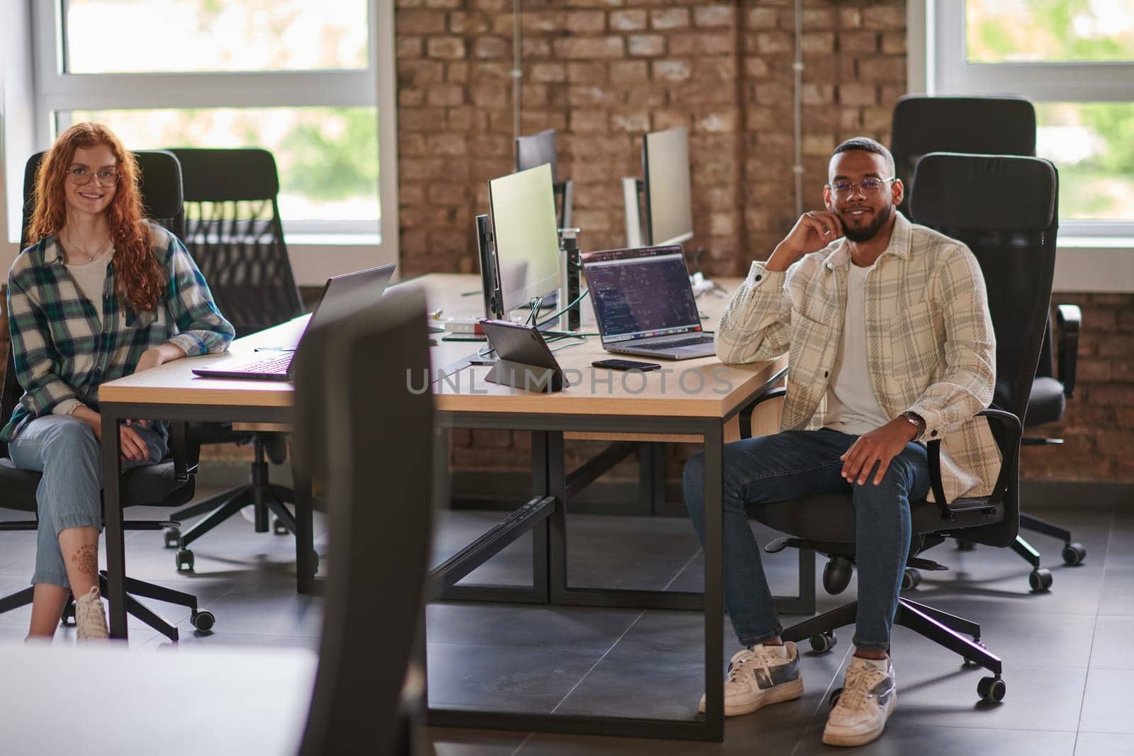 Group of colleagues, a woman with vibrant orange hair and a young African American businessman, sitting in a modern office space, symbolizing diverse collaboration and a dynamic work environment