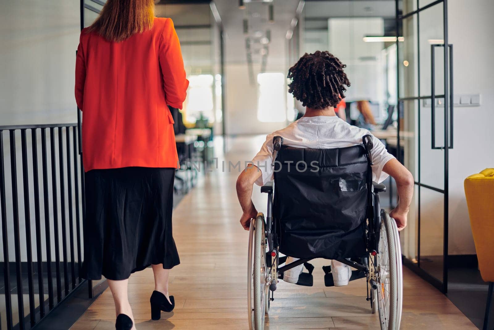 A group of young business people in a modern glass-walled office captures the essence of diversity and collaboration, while two colleagues, including an African American businessman in a wheelchair, reflect inclusivity and discuss solving business problems.