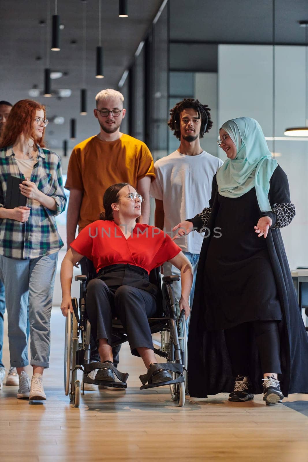 A diverse group of young business people walking a corridor in the glass-enclosed office of a modern startup, including a person in a wheelchair and a woman wearing a hijab, showing a dynamic mix of innovation and unity. by dotshock