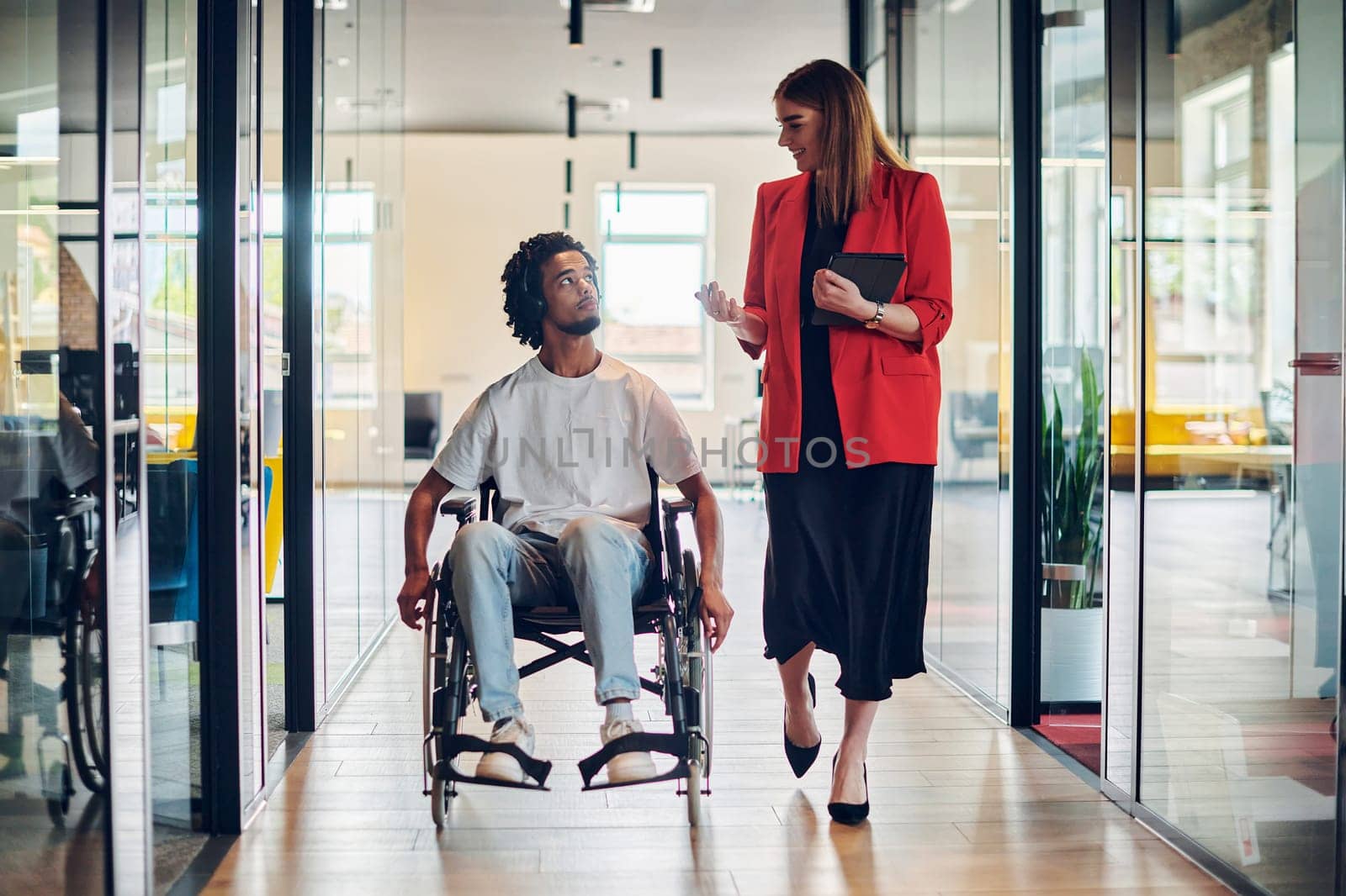 A group of young business people in a modern glass-walled office captures the essence of diversity and collaboration, while two colleagues, including an African American businessman in a wheelchair, reflect inclusivity and discuss solving business problems.