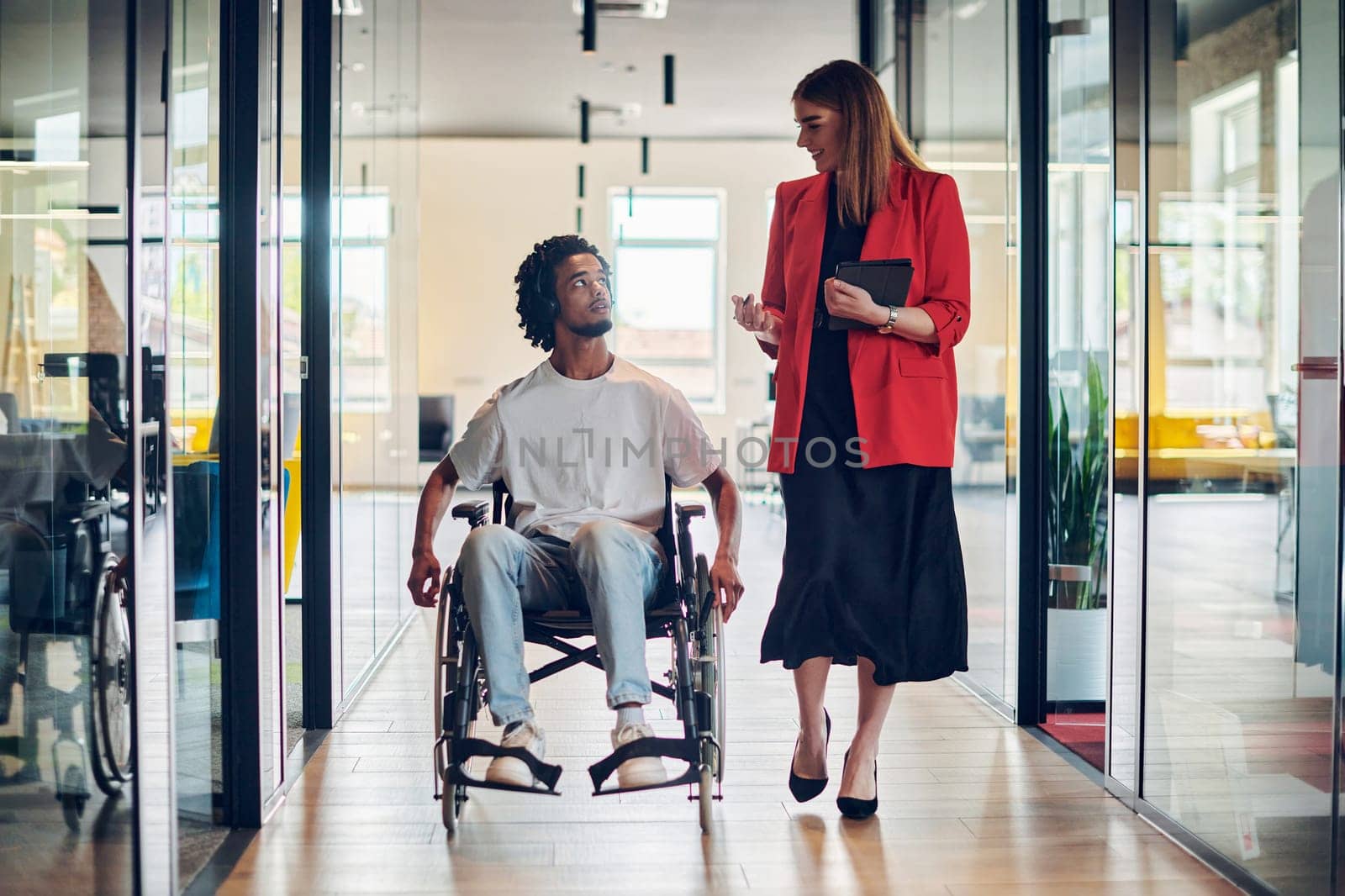 A group of young business people in a modern glass-walled office captures the essence of diversity and collaboration, while two colleagues, including an African American businessman in a wheelchair, reflect inclusivity and discuss solving business problems.