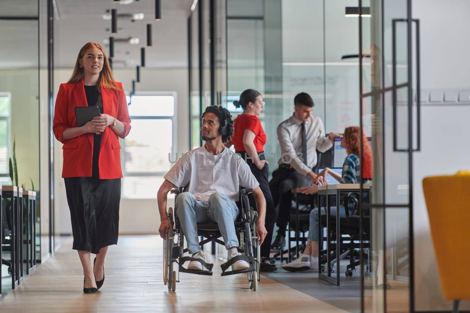 A group of young business people in a modern glass-walled office captures the essence of diversity and collaboration, while two colleagues, including an African American businessman in a wheelchair, reflect inclusivity and discuss solving business problems.