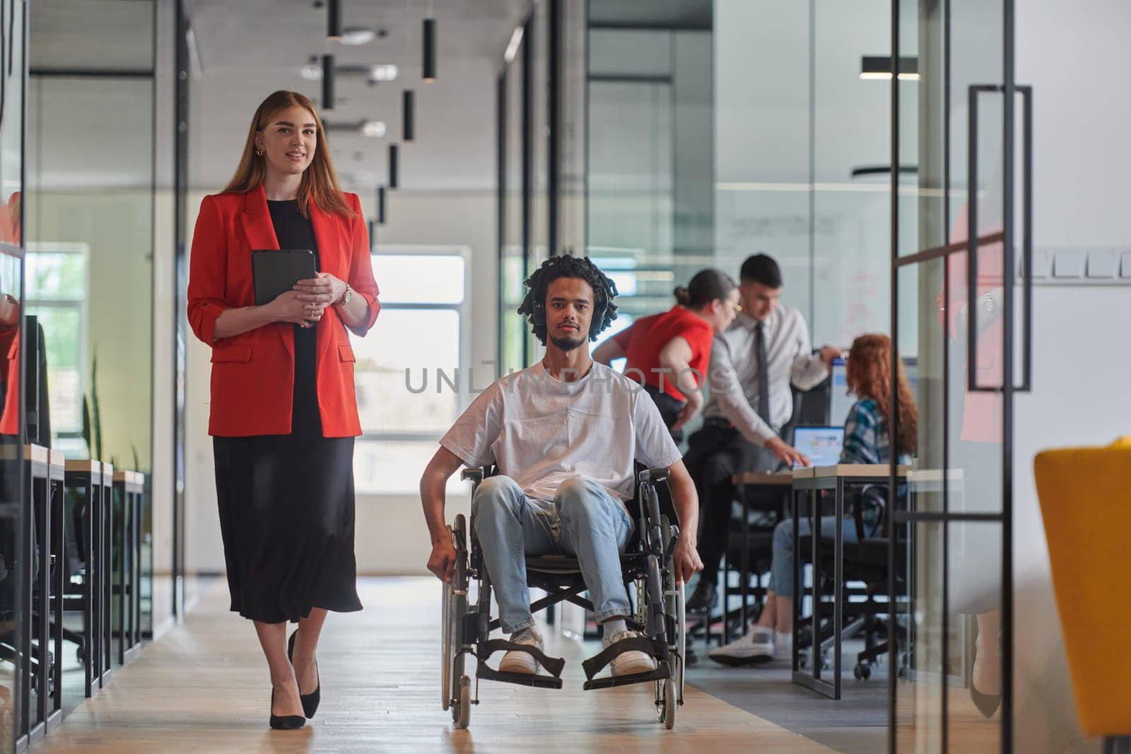 A group of young business people in a modern glass-walled office captures the essence of diversity and collaboration, while two colleagues, including an African American businessman in a wheelchair, reflect inclusivity and discuss solving business problems.