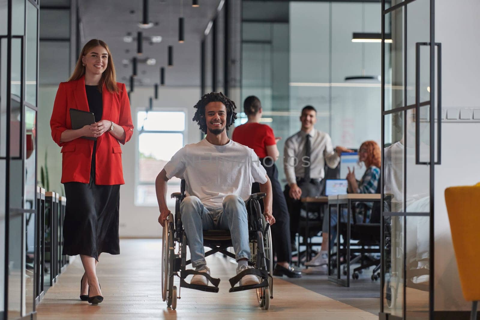 A group of young business people in a modern glass-walled office captures the essence of diversity and collaboration, while two colleagues, including an African American businessman in a wheelchair, reflect inclusivity and discuss solving business problems.