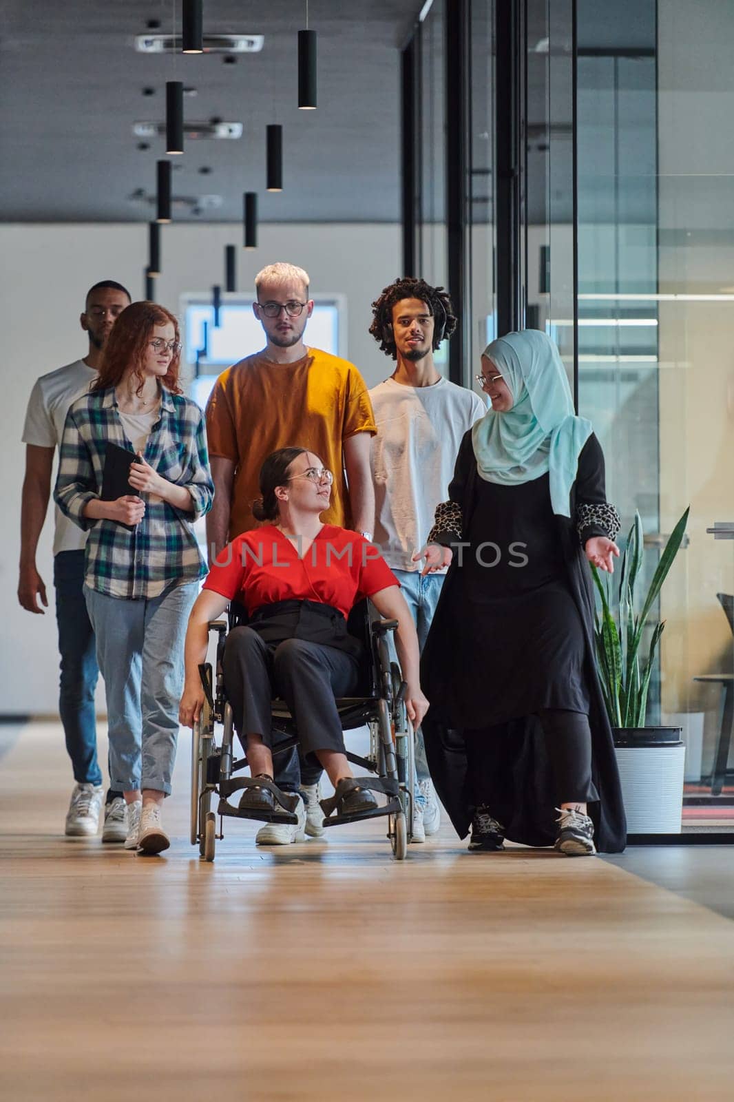 A diverse group of young business people walking a corridor in the glass-enclosed office of a modern startup, including a person in a wheelchair and a woman wearing a hijab, showing a dynamic mix of innovation and unity