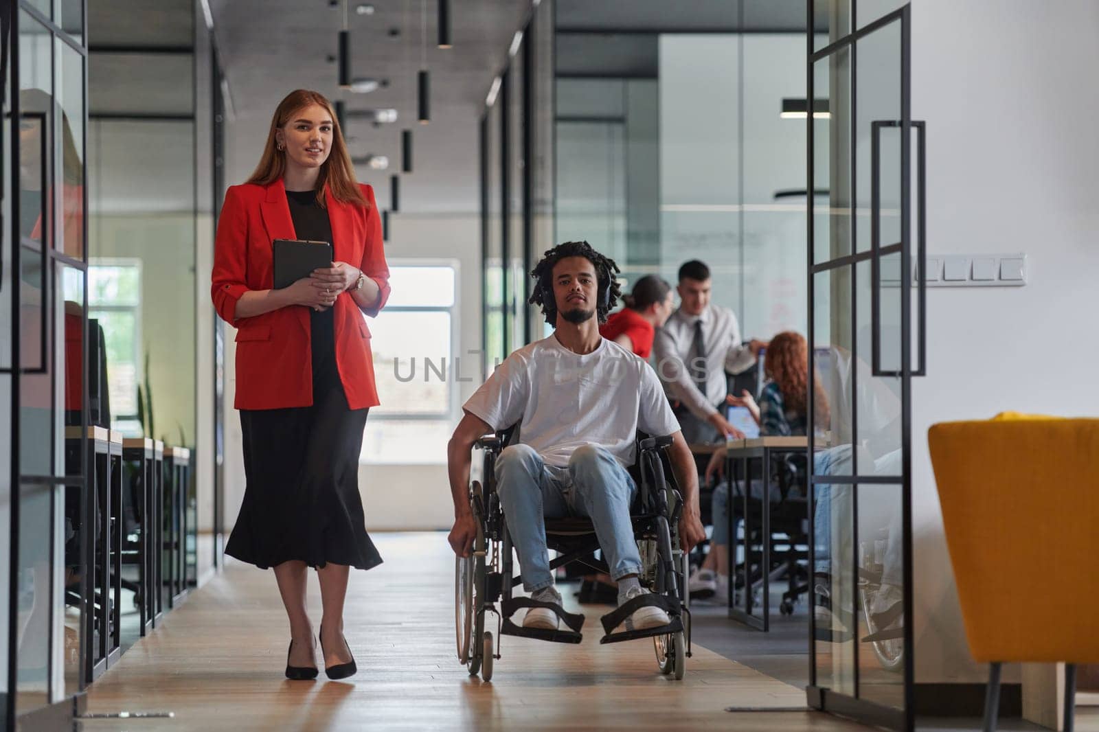 A group of young business people in a modern glass-walled office captures the essence of diversity and collaboration, while two colleagues, including an African American businessman in a wheelchair, reflect inclusivity and discuss solving business problems.