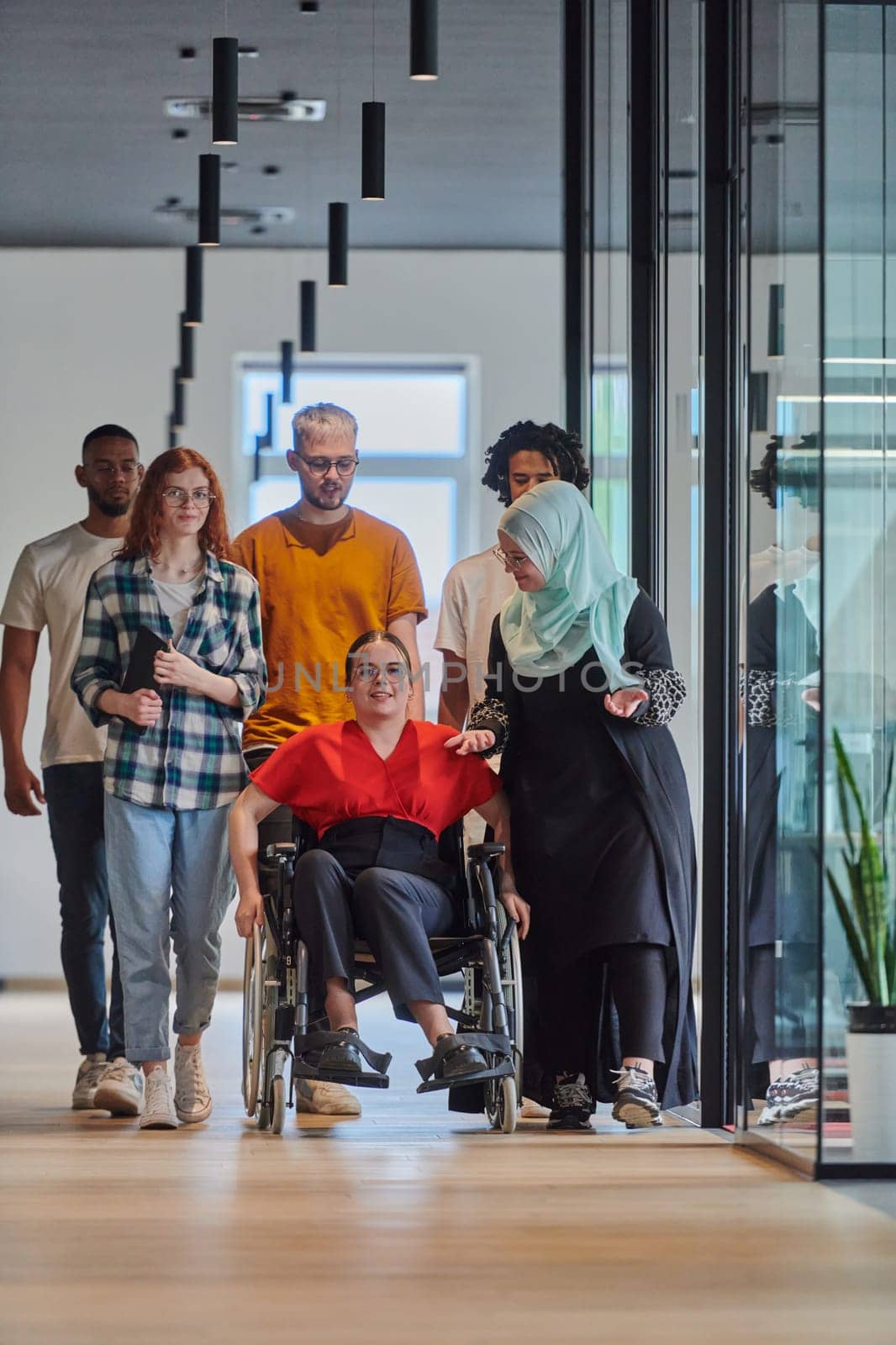 A diverse group of young business people walking a corridor in the glass-enclosed office of a modern startup, including a person in a wheelchair and a woman wearing a hijab, showing a dynamic mix of innovation and unity