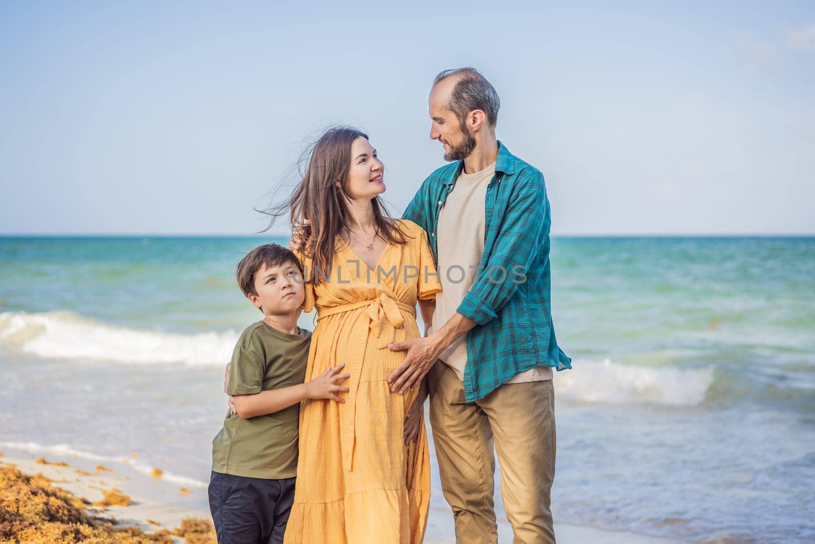 A loving family enjoying tropical beach - a radiant pregnant woman after 40, embraced by her husband, and accompanied by their adult teenage son, savoring precious moments together amidst nature's beauty. Pregnancy after 40 concept by galitskaya