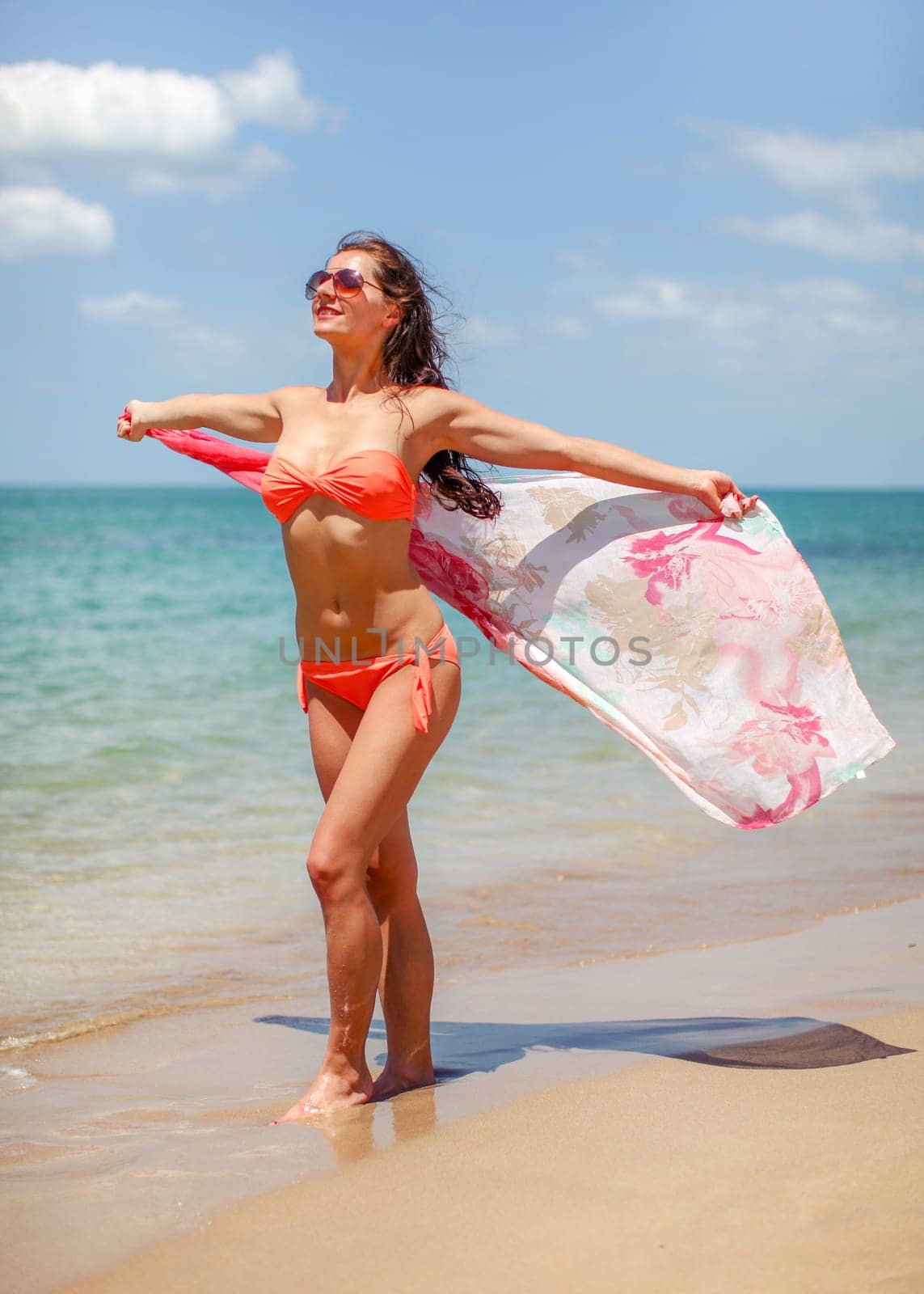 Young woman wearing orange red bikini and sunglasses standing on the beach waving silk scarf in wind behind her, sea background. by Ivanko