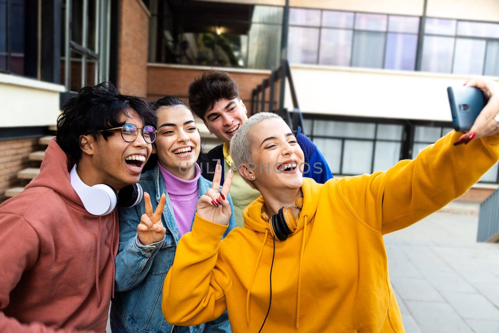 Happy multiracial college student friends take selfie with mobile phone laughing and having fun outside university building. Social media concept.