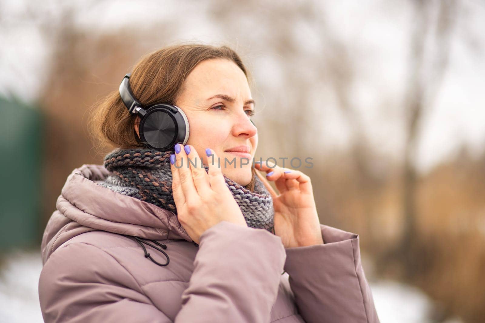 Joyful young woman in jacket listening to music via black headphones, relaxing in a park