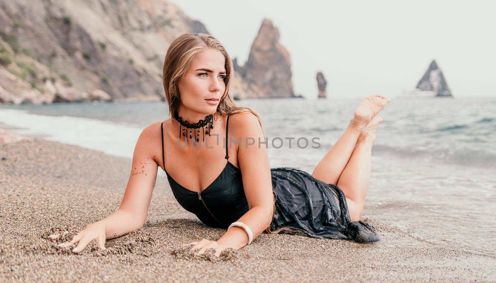 Woman summer travel sea. Happy tourist in black dress enjoy taking picture outdoors for memories. Woman traveler posing on sea beach surrounded by volcanic mountains, sharing travel adventure journey by panophotograph