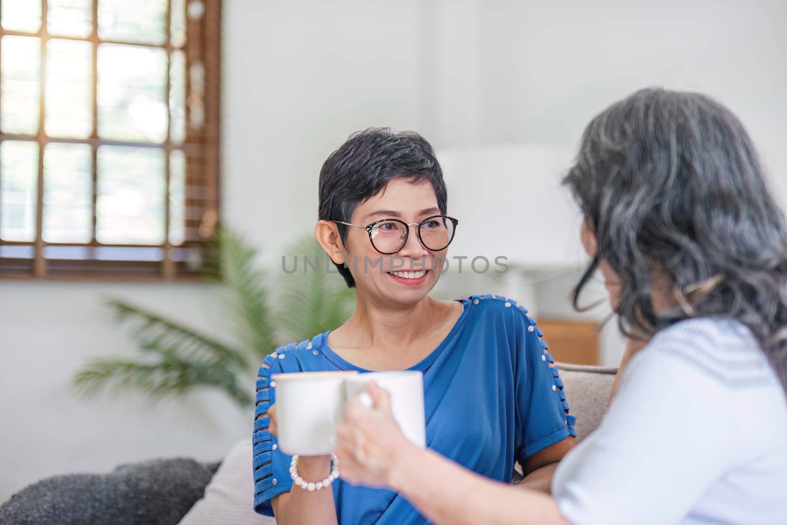 Two Asian women are sitting drinking coffee and chatting in the living room. by wichayada